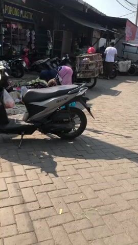 A woman wearing a headscarf and pink pants is bending over, sorting through items on the ground. In the foreground, a silver scooter is parked on a paved walkway. Behind the woman, a store with the words "PORONG PLASTIK" is visible. Several other motorcycles are parked nearby, and a caged item, possibly containing live animals, is attached to the back of a motorcycle. Another person, partially visible, is standing to the right. The environment appears to be an outdoor market or street.