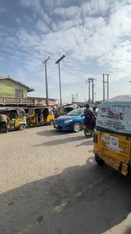 The video shows a street scene with several vehicles and people. In the foreground, a blue sedan is driving down a paved road. To the right, the back of a yellow auto-rickshaw is visible, with some writing on its side. A man wearing a yellow shirt and denim shorts walks from left to right in the middle ground. Behind the blue car, several yellow auto-rickshaws are parked. Further in the background, there are buildings with shops and people. A motorcycle with two riders passes by in the middle ground. The sky is partly cloudy. The overall impression is a busy street in a town or city.