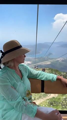 This media file shows a woman in a cable car ascending Mount Lovćen in Kuk, Montenegro. The woman is wearing a straw hat with a black ribbon and a light blue, sheer blouse with floral embroidery on the sleeves. She is seated and looking out the window at the stunning panoramic view. The cable car has tan seats and black trim, and a logo that reads "KOTOR CABLE CAR" is visible on the window.

The scene outside the cable car is a vast landscape of mountains and the Bay of Kotor. The water is a deep blue, and the distant mountains are shrouded in a light haze, suggesting a warm, clear day. The sun is high in the sky, casting bright light and creating distinct shadows, indicating it is likely midday. There are other cable cars visible in the distance, also ascending the mountain. The foreground shows rugged, green terrain, typical of a mountainous region. The woman's posture and gaze suggest a sense of peaceful contemplation and enjoyment of the breathtaking scenery. The overall impression is one of a tourist enjoying a scenic ride and the magnificent natural beauty of the Montenegrin coast.