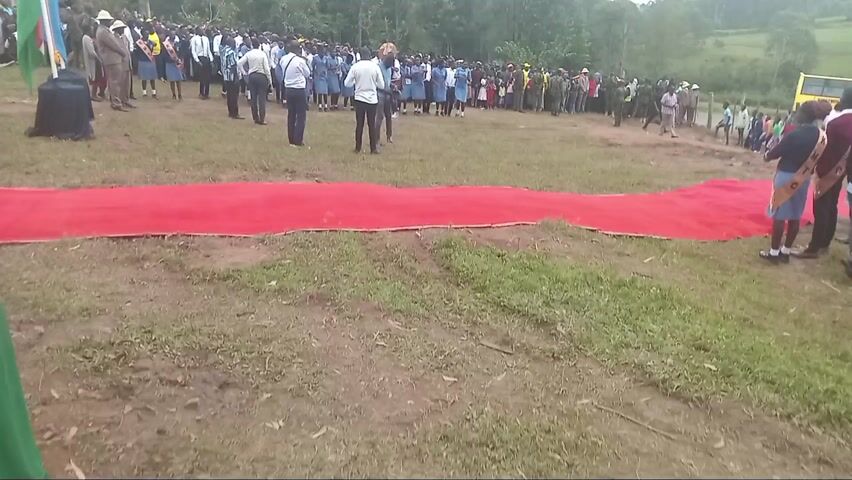 The video shows a large outdoor gathering in a grassy field in Moticho ward, Kenya, on an overcast day. A significant crowd of people, primarily young students, are assembled. Many students are dressed in uniforms, with some wearing white shirts and red ties, and others in light blue dresses or uniforms. Several adults are also present, including some in official-looking uniforms (possibly military or police) and others in civilian attire.

A long red carpet is laid out on the ground, extending from the bottom center towards the right. On the far left, two flags are visible next to a black draped table; one appears to be the Kenyan national flag, and the other features yellow, green, and blue colors.

In the foreground, several adults are positioned, some holding cameras or phones, indicating they are recording or photographing the event. Towards the center-right of the assembled group, an individual holds a round, decorated object that resembles a traditional drum. Among the students on the left, one young woman is wearing a yellow sash with the partial text "KM... TO" visible on it. The background features a dense cluster of trees, suggesting a rural or semi-rural setting. The overall scene depicts a formal or ceremonial assembly, possibly a school event or community gathering, with participants appearing attentive.