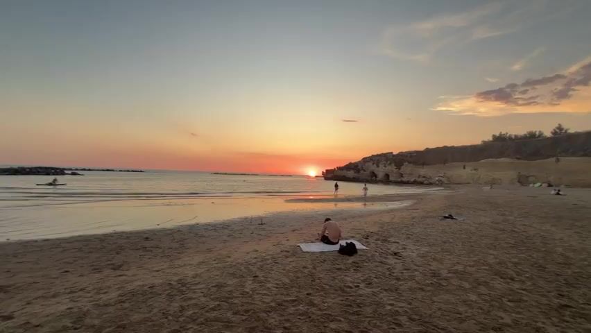 A wide, static shot captures a serene beach at sunset. The sun is low on the horizon, casting a warm orange glow across the sky and reflecting on the calm ocean. The beach is sandy, with some scattered rocks and a rocky cliff face to the right. Several people are visible. In the foreground, a person sits on a towel facing the ocean. Further down the beach, two figures stand near the water's edge. In the distance, on the water, a person is in a kayak. The overall atmosphere is peaceful and tranquil.