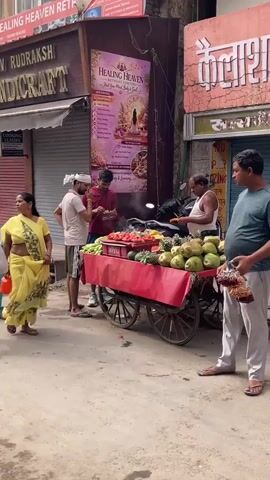 The video shows a street scene with a fruit cart in the center, laden with various fruits like watermelons, pineapples, and bananas. Two women, one in a yellow sari and another in a light green kurta, are walking past the cart. A man in a blue t-shirt and grey pants is standing on the right side of the cart, facing it and appearing to interact with the vendor. Another man, presumably the vendor, wearing a light blue shirt and grey trousers, stands behind the cart, handing something to the man in blue. Several other people are visible in the background, including a woman in a teal outfit walking towards the cart. The setting appears to be a market or a busy street, with shops and signs in the background. The weather seems to be daytime with natural lighting.