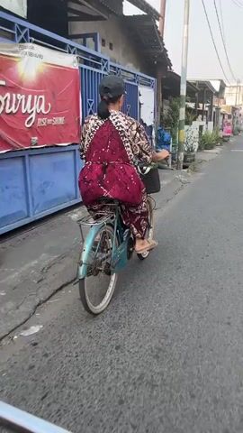 A person is riding a bicycle down a paved street. The cyclist is wearing a long, patterned dress and a scarf. A basket is attached to the front of the bicycle. The street is lined with houses and shops. There are other people and vehicles in the distance. The sun is shining, casting shadows on the street.
