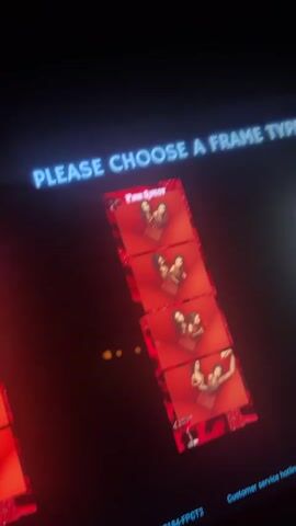 A photobooth screen displays a selection of photo frame templates. The screen is angled and partially obscured by darkness. The top of the screen has text that reads "PLEASE CHOOSE A FRAME TYPE" in a bright blue font. Below the text is a vertical strip of four identical photo frames. Each frame shows a close-up of two women posing. The background of the frames is red with a glittery pattern, and the borders have a black and red leopard print design. In the lower right corner of the screen, there is a line of text that reads "Customer service hotline:+1". In the lower left corner, there is another line of text with a store ID. The scene is lit by the screen itself, casting a red and blue glow on the surrounding dark environment. A few small yellow spheres are visible in the lower left quadrant of the screen, possibly a reflection or part of the display.