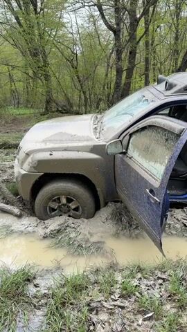 A blue Nissan Xterra SUV is deeply stuck in a large mud pit in a forest. The vehicle is heavily soiled with mud splatters across its body, windows, and roof rack. The front driver's side door is open, revealing a muddy interior. The SUV's tires are completely submerged in the thick, light brown mud and standing water. The surrounding ground is also muddy, with patches of green grass and fallen leaves. In the background, numerous bare and lightly foliated trees indicate a wooded setting, possibly during spring. The light suggests it is daytime.