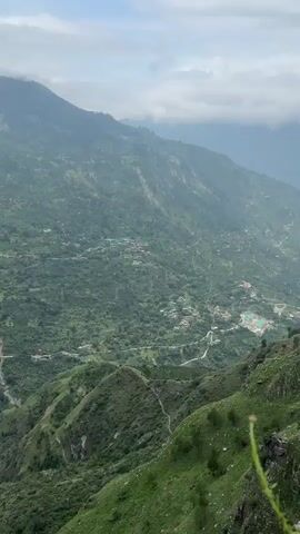 The video shows a panoramic view of a mountainous landscape with lush green vegetation covering the slopes. Small clusters of buildings, likely houses and possibly a small village, are scattered across the hillsides. Several power lines and transmission towers are visible, indicating some level of infrastructure in the area. Winding roads or paths can be seen snaking through the terrain, connecting the scattered settlements. The sky appears overcast with clouds. The camera is positioned at a high vantage point, looking down into the valley and across the rugged terrain. The overall scene depicts a rural, mountainous region.