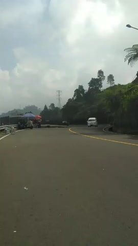 The video shows a road in a mountainous area with traffic.  A white van rounds a curve in the road, followed by several motorcycles. The motorcyclists, wearing helmets, are riding on the right side of the road.  On the left side of the road, near the camera, there are some street vendor stalls set up under umbrellas. A green hillside covered with trees rises on one side of the road, and across the valley on the other side is another hillside and electrical towers.  The sky is overcast.