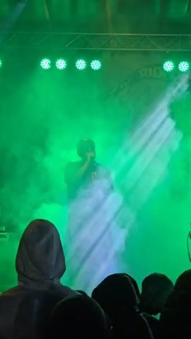 A low-angle, handheld video shows a rapper performing on a stage full of green smoke. The stage features an overhead rig of round LED lights and a sign behind the performer that reads “The Riders Crew.” The rapper wears a cap, t-shirt, and shorts. He stands in front of a microphone and raps to the audience. A beam of light periodically flashes down from the rig. The audience is visible in the foreground, mostly showing the backs of heads and the hoods of clothing.