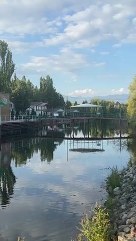 The video shows a calm body of water with a bridge spanning across it. The bridge is red and has a white canopy on top. Trees and buildings are visible on the shore behind the bridge. The sky is blue with some clouds, and mountains can be seen in the distance. The water reflects the bridge, trees, and sky. The scene appears to be a park or a resort.