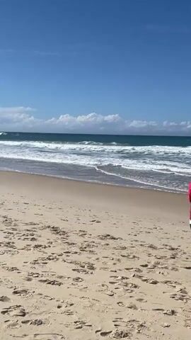 A woman wearing a red quilted jacket and jeans walks away from the camera towards the ocean on a sandy beach. The ocean is blue with white waves breaking on the shore. The sky is clear blue with a few white clouds. Another person, possibly a man, is standing in the shallow water wearing a red hat and shorts, facing the ocean.