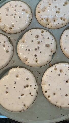 The video shows a close-up of a muffin tin filled with pancake batter. The batter is light in color and speckled with small dark dots. Bubbles are forming on the surface of the batter as it cooks. The muffin tin appears to be made of a gray metal material.