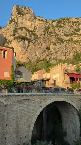 The video shows a view of a village in front of a rocky mountain. The village has stone buildings with terracotta roofs. A stone bridge with an arch spans over a stream or waterfall. There are plants and flowers on the bridge and balconies of the buildings. The lighting suggests it is either sunrise or sunset, with warm golden light illuminating the scene. There are no people visible in the video.
