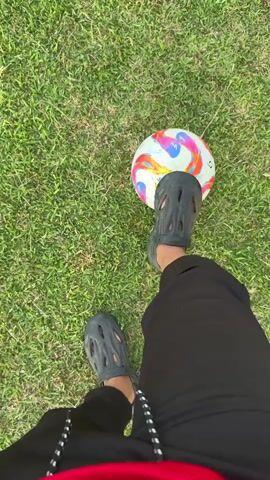 The video shows a person's legs and feet, wearing black Crocs and black pants, standing on a green grassy field. A colorful soccer ball is placed in front of the person's right foot. The person then kicks the soccer ball, and it rolls away. The scene appears to be outdoors, with sunlight indicating it's daytime. The grass is well-maintained, suggesting a park or sports field. The overall atmosphere is casual and recreational, with no indication of a formal event. There is no visible text in the video. The action is simple and direct, focusing on the act of kicking a soccer ball.