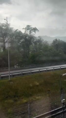 The video shows a view of a wet highway from an elevated position, possibly from a passing vehicle. The weather is overcast and it appears to be raining or hailing. Green grass and trees line the highway's edge. A red convertible car drives along the highway. In the background, mountains are visible through the mist. There is an indistinct voice, seemingly that of a child, heard in the background.