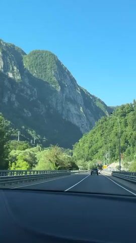 This is a first-person perspective video recorded from inside a vehicle, likely a car, as it travels along a highway. The highway cuts through a scenic mountain valley. The mountains are steep and covered in lush green vegetation, with some exposed rock faces. The sky is a clear, bright blue, suggesting fair weather and a daytime recording.

The highway itself has multiple lanes and is lined with guardrails. There are a few other vehicles visible on the road, including cars and motorcycles, indicating ongoing traffic. Streetlights and utility poles are also present along the roadside. In the distance, what appears to be a tunnel entrance is visible on the right side of the road.

The overall mood of the video is one of travel and appreciation for natural beauty. The setting is likely a mountainous region in Russia, given the context provided. There is no discernible text within the video frame itself. The time of day is clearly daytime, and the weather appears to be sunny and pleasant.