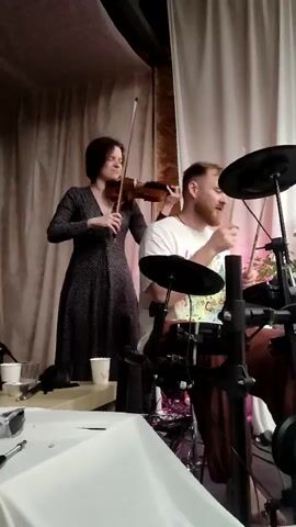 The video begins with a close-up of a drummer playing an electronic drum set, with a woman playing the violin behind him. The camera then pans to a bassist playing a red electric bass guitar, and a singer holding a microphone. The scene shifts again to show a guitarist playing an acoustic guitar, a keyboardist, and a drummer playing a djembe. The musicians appear to be performing in an indoor venue with draped fabric and plants visible in the background.