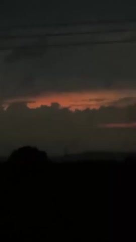 A static shot captures a view of a dusk or dawn sky. The sky is predominantly grey, with a strip of orange light visible through the clouds near the horizon. The silhouettes of trees or bushes are visible in the foreground, obscuring the ground. Utility poles and power lines are visible against the sky.