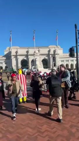 A group of people march down a brick-paved walkway in front of a large, ornate building with three American flags on its roof. The marchers carry a banner that reads "NO FASCIST AMERICA GET 2 DO." Many of the marchers are dressed in red cloaks and white bonnets, reminiscent of the costumes from "The Handmaid's Tale." Several people are taking photos or videos of the march. The sky is clear and blue. In the background, there is a traffic light and a few other people walking by.