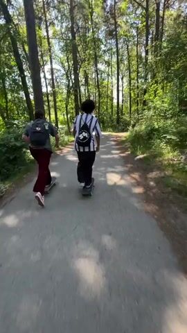The video shows two young people skateboarding on a paved path in a wooded area. The person in the foreground is wearing a dark t-shirt, maroon pants, and a backpack. They are skateboarding from left to right in the frame. The second person, further ahead and to the right, is wearing a striped shirt and dark pants, also with a backpack. They are also skateboarding away from the camera. The path is surrounded by trees with green foliage, and sunlight filters through the leaves, creating dappled shadows on the ground. The overall impression is of a leisurely activity in a natural setting.