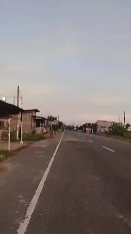 The video shows a road with buildings on the side. The road is asphalt and has white lines painted on it. There are some potholes in the road. The sky is overcast. The buildings appear to be a mix of residential and commercial. There are trees and vegetation along the side of the road. The video appears to be taken from a moving vehicle.