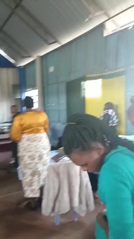 The video begins with a close-up of a woman's braided hair in a teal-colored hooded sweatshirt. The camera then pans to reveal a group of people in what appears to be a gathering or service. A woman in a bright pink dress stands out, with her hair in an updo. To her right, a man in a black shirt is partially visible. In the background, a child with braided hair is visible. The environment seems to be an indoor space with corrugated metal walls and a simple ceiling. The camera shifts again to show a wider view of a gathering, possibly a church or a similar community event. People are dressed in various outfits, including a woman in a bright orange garment and a woman in a patterned skirt and yellow top. Some individuals are standing, while others are seated. There is a raised platform or altar area draped with blue and white fabric, and what appears to be a casket is visible on the platform. The floor is a reddish-brown color. The lighting suggests it is daytime, with light coming from windows. The atmosphere seems somber, given the presence of a casket, but there are also signs of community interaction.