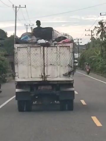 A video taken from inside a moving vehicle shows the rear view of a white dump truck from a slightly elevated perspective. The truck's bed is overflowing with various items, appearing to be trash or salvaged goods. A silhouette of a person is visible sitting atop the pile of items in the truck bed, facing away from the camera. The truck is driving on a paved road with a solid white line on the left and a broken white line on the right, indicating two lanes of traffic. Utility poles and wires line the left side of the road, and trees and foliage are visible on both sides. Another white vehicle is visible in the distance on the right side of the road, and a motorcycle is partially visible on the far right. The sky is overcast.
