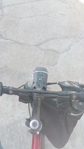 A first-person perspective video shows a person riding a bicycle on a paved road. The camera is mounted on the handlebars. In the foreground, a red cycling computer with a digital display is visible on the left handlebar. A black bicycle light is mounted in the center of the handlebars, above the stem. A black bag is attached to the handlebars or stem. The road surface appears worn and cracked, with tire tracks visible in places. In the background, there are buildings, cars, and trees. The sun is shining, casting shadows on the road. The video captures the motion of the bicycle as it travels forward.