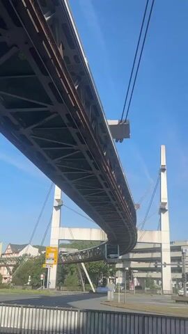 The video shows a monorail train traveling on a track suspended above a street. The train is blue and white and has the number 01 visible on its side. The track is a steel structure supported by white pylons. There are buildings and trees visible in the background, and the sky is blue with some clouds. The camera appears to be looking up at the monorail from below. The video is filmed from a moving vehicle, likely another car or bus, as indicated by the slight motion blur and the railing at the bottom of the frame. The monorail appears to be in motion, as the train is clearly visible under the track. There are no people clearly visible in the video.