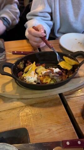 Two individuals wearing grey sweatshirts sit at a wooden table, sharing a skillet of nachos. One person uses a fork and their hands to eat the nachos. The skillet sits on a round wooden board. The table also contains a plate with a single nacho, cutlery, and a beverage.