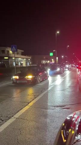 The video captures a night-time street scene, likely a residential or commercial area, with several cars driving in the same direction. The road surface appears cracked and illuminated by streetlights and the headlights of the vehicles. In the foreground, the rear of a dark-colored car is visible, with its red taillights illuminated. Further down the road, a line of cars is moving, including a dark red sports car on the left, a blue sports car in the middle, and several other vehicles behind them. The red sports car appears to be performing a burnout, as indicated by smoke emanating from its rear tires. The blue sports car has its headlights on, casting a bright beam onto the road. Buildings are visible on the left side of the street, with illuminated windows suggesting they are businesses or residences. A pedestrian crossing sign is also present. The overall atmosphere is that of a casual car gathering or street cruising event at night.