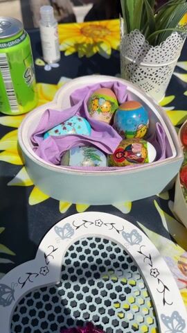 The camera pans across a table set for an outdoor gathering. A heart-shaped box filled with Easter eggs rests on a sunflower-patterned tablecloth. Next to it, a wooden container holds fresh strawberries. An egg carton filled with speckled eggs sits further back, along with a cupcake decorated with a chocolate egg. The camera movement reveals more of the tablecloth, which features a bee design in addition to sunflowers.