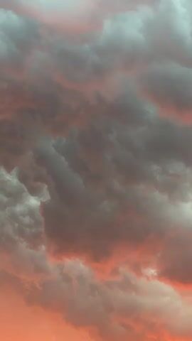 This is a close-up, vertical video of a cloudy sky during sunset. The clouds are a mix of dark gray and reddish-orange, with some lighter, wispy clouds interspersed. The light suggests the sun is low on the horizon, casting a warm glow on the clouds. The camera is stationary, and there are no discernible objects, people, or actions within the frame. The environment is solely the sky and its atmospheric conditions.