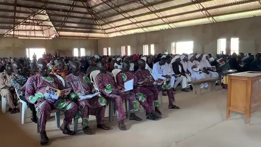 This media file depicts an indoor gathering of people, likely a religious service or a community event, taking place in a large, open hall with a corrugated metal roof. The walls are unpainted concrete, and natural light streams in through numerous rectangular windows spaced along the length of the building.

The subjects are primarily individuals dressed in traditional African attire. In the foreground, four men are seated in chairs, wearing matching vibrant, patterned garments with green accents and red or maroon bases. They are holding papers or programs. Behind them, the hall is filled with a diverse crowd of people, many wearing white or light-colored clothing, suggesting a formal or religious occasion. Some individuals in the back appear to be holding musical instruments, possibly drums, hinting at a musical component to the event.

Objects visible include several wooden benches, a wooden podium or cabinet in the foreground, and at least two drums further back in the hall. The floor is earthen. The overall impression is one of a communal gathering, possibly in a rural or semi-rural setting given the simple construction of the building. The lighting suggests it is daytime, and the interior is somewhat dusty, hinting at a dry climate.

The expressions and postures of the individuals in the foreground suggest attentiveness, with some looking forward at an unseen focal point, while others are engaged with their papers. The presence of a large gathering in a place of worship or community hall indicates a significant event is taking place. There is no discernible text visible in the image. The location context provided is Kufana, Nigeria.