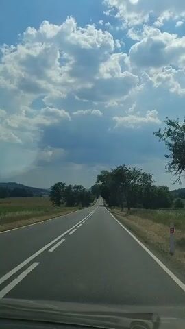 The video shows a view from a car traveling on a paved road through a rural landscape. The road has a white dividing line. On either side of the road are fields and trees. The sky is blue with fluffy white clouds. The time of day appears to be daytime, with sunlight casting shadows. The weather seems to be fair. The scene evokes a feeling of a peaceful journey through the countryside. No text is visible in the video.