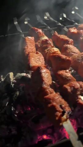 The video shows skewers of meat being grilled over hot coals at night. Smoke rises from the grill, and in the background, distant lights suggest an urban or suburban setting. The meat appears to be seasoned and is developing a browned, slightly charred exterior. There are onions interspersed with the meat on some of the skewers. The camera angle is close to the grill, focusing on the food. The scene is dark, lit primarily by the glow of the coals and the food itself. The audio includes snippets of conversation in Russian.