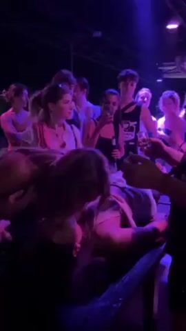 A group of people are gathered in a dimly lit space, illuminated with occasional flashes of blue and purple light. They appear to be at a party or gathering, some holding drinks. In the foreground, a person is lying on what looks like a table or platform, while others stand around them. Some of the people are clapping and raising their hands in the air, seemingly celebrating. The overall atmosphere suggests a festive and lively event.