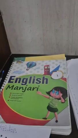 The video shows a book titled "English Manjari" with the number 1 on it. The book is green and red and has a cartoon character on the cover. There are also some papers scattered around the book. The background appears to be a wall with some writing on it. The video then shifts to show a row of empty chairs and desks in what appears to be a classroom or office setting. The lighting is dim and the overall impression is one of disuse or emptiness.