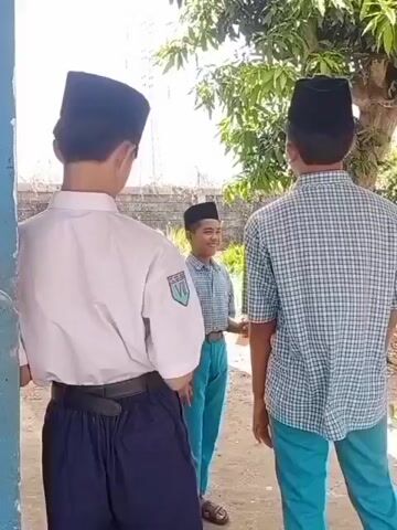The video shows four young males outdoors, seemingly in a schoolyard or similar setting. The background includes trees, buildings, and a paved walkway. Three of the males are wearing traditional black skullcaps, known as peci or songkok, and school uniforms consisting of plaid shirts and teal pants. One male is wearing a white shirt and dark pants. The first scene focuses on two males facing each other, with a third male smiling behind them. The camera then pans to show one of the males in the plaid shirt turning and looking at the camera. The video then shows two males in plaid shirts and teal pants interacting near a blue pillar. One of them is holding a drink in a small pouch and appears to be drinking from it, while the other stands beside him. The lighting suggests it is daytime.