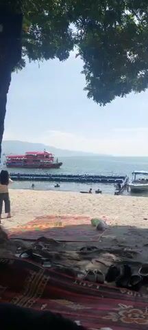 The video shows a sandy beach with a body of water in the background. A red passenger boat is visible on the water, as are several smaller boats and what appear to be floating structures. There are a few people visible in the water and on the beach. The scene is framed by tree branches at the top of the video. The weather appears to be sunny.
