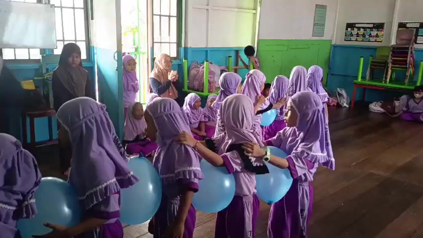 A group of young girls in purple uniforms and headscarves are participating in an activity in a classroom. Some of the girls are holding light blue balloons between their knees as they stand in a line, linking arms. A woman wearing a headscarf and holding a microphone is standing on the left side of the frame, facing the girls. She appears to be addressing them or giving instructions. In the background, other children and adults are visible, and the room appears to be a classroom with wooden floors and colorful walls. There are also stacks of paper cups on a table on the left side of the frame. The overall scene suggests a children's event or game.