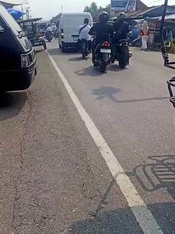 A street scene, likely in a developing country. Several vehicles are on the road, including a black van on the left, motorcycles, and a three-wheeled vehicle. In the background, there are buildings that appear to be shops. People are also visible, some riding on motorcycles and others standing. The road is paved with asphalt and has a white line dividing the lanes. The lighting suggests it is daytime, possibly midday, with shadows cast by the vehicles and the sun. The overall impression is a busy street with moderate traffic.