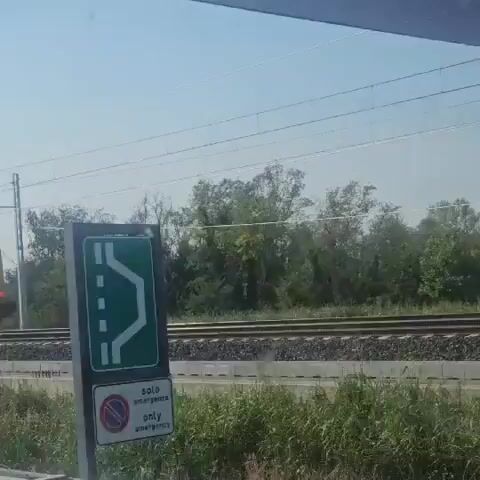 A video captures a yellow train moving on railway tracks. The train appears to be carrying equipment or materials. The surrounding environment consists of trees and vegetation, with a clear sky overhead. The perspective suggests the video was filmed from a moving vehicle, likely a car, looking through the windshield. The train is the central focus, and the motion implies a continuous journey.