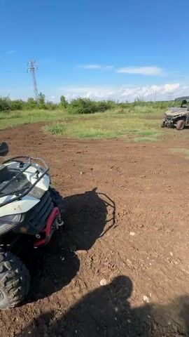 The video shows a dirt area with two ATVs parked. One ATV is in the foreground and the other is further back, slightly to the left. The ATVs are black and appear to be muddy. There is also a UTV in the background, parked to the left of the ATVs. The area is a field with dry grass and some bushes. The sky is blue with some white clouds. The video is taken from a low angle.