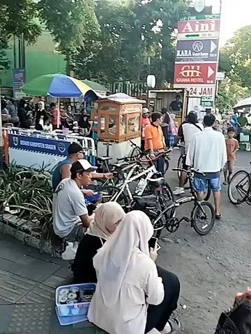 A daytime outdoor shot depicts a bustling street scene filled with people, bicycles, and market stalls. In the foreground, three individuals are seated on a curb, two women in hijabs using phones, and a man in a white cap and t-shirt with two other men. A pile of bicycles is parked nearby. To their right, more bicycles are scattered, some with riders, and many pedestrians are visible, moving around or standing. Market stalls with colorful umbrellas and goods are set up along the street. In the background, buildings and trees line the road. A large sign with "AINI" and "24 JAM" is visible on the right side. The overall atmosphere appears lively and busy.