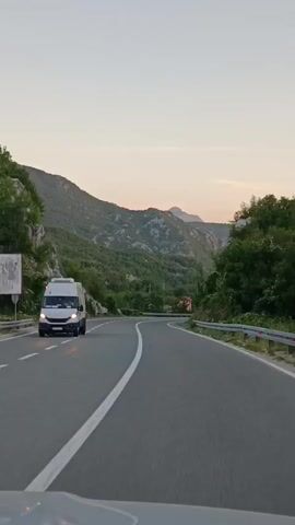 A car is driving on a winding road through a mountainous, forested area. The sky is clear and light, suggesting it is either sunrise or sunset. The road has a white line separating the lanes. On the left side of the road, there is a rocky cliff face and dense green vegetation. On the right side, there is a metal guardrail and more trees. A white van is visible in the distance on the left side of the road.