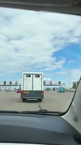 A white camper van with a drawing of a face and the text "your Heart is the Light of this world" written on the back is seen from behind driving through a toll plaza on a sunny day. Several other cars are also in line at the toll plaza. The sky is blue with white clouds.
