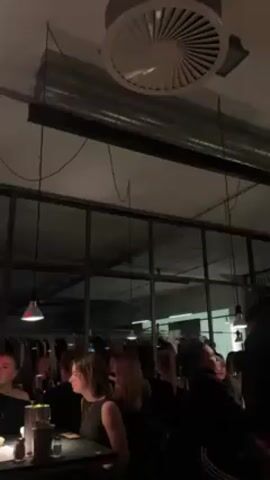 The video shows a dimly lit, crowded interior space, possibly a bar or clothing store. Several people are standing and sitting, mostly in dark clothing, engaged in conversations. Some are holding drinks. In the background, there are racks of clothing and a large window. Overhead, a circular ventilation fan is visible, along with exposed pipes and wires. The lighting is provided by several small hanging lamps.
