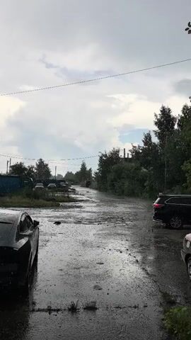 A black SUV is driving on a wet, rain-slicked road. The sky is cloudy and it is raining. There are other cars parked on the side of the road and trees in the background. The road appears to be in a rural or industrial area.