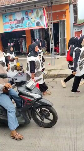 A group of people are walking down a street, some are wearing cow costumes. A few people are riding on motorcycles, and one person is pushing a stroller. There are buildings on either side of the street, and a flag is visible on one of the buildings. The weather appears to be overcast.
