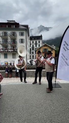Three men are performing music outdoors. The man on the left is playing a sousaphone. The man in the center is playing a banjo. The man on the right is playing a clarinet. They are all wearing vests and hats. Behind them are buildings and mountains. A white banner with text is to the right of the musicians.